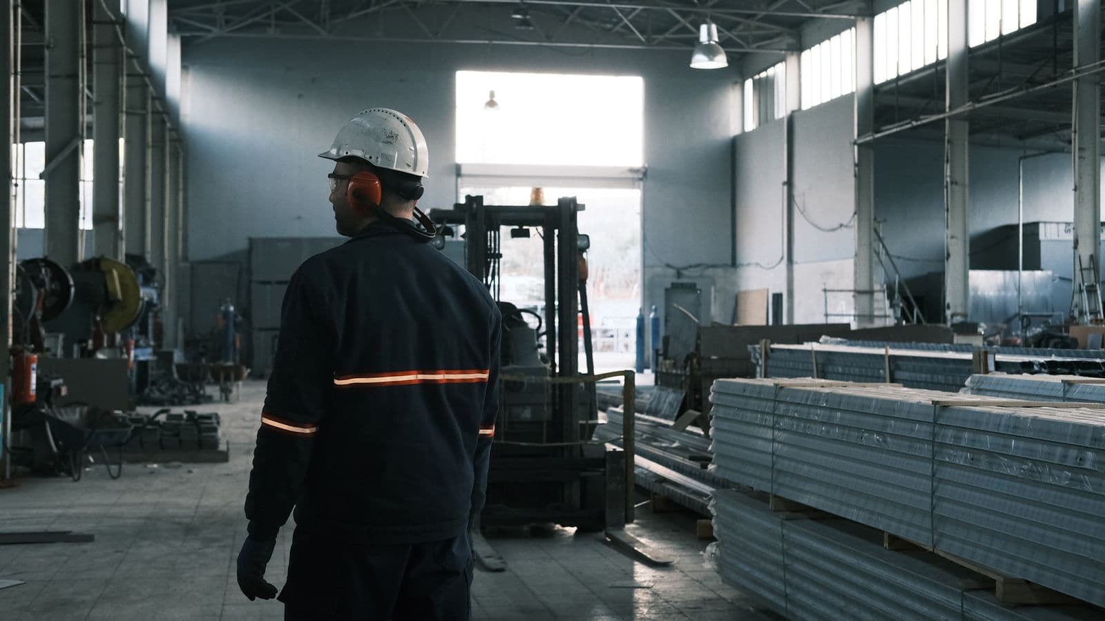 Trabalhador de costas observando a operação em um galpão industrial, com material empilhado e empilhadeira ao fundo