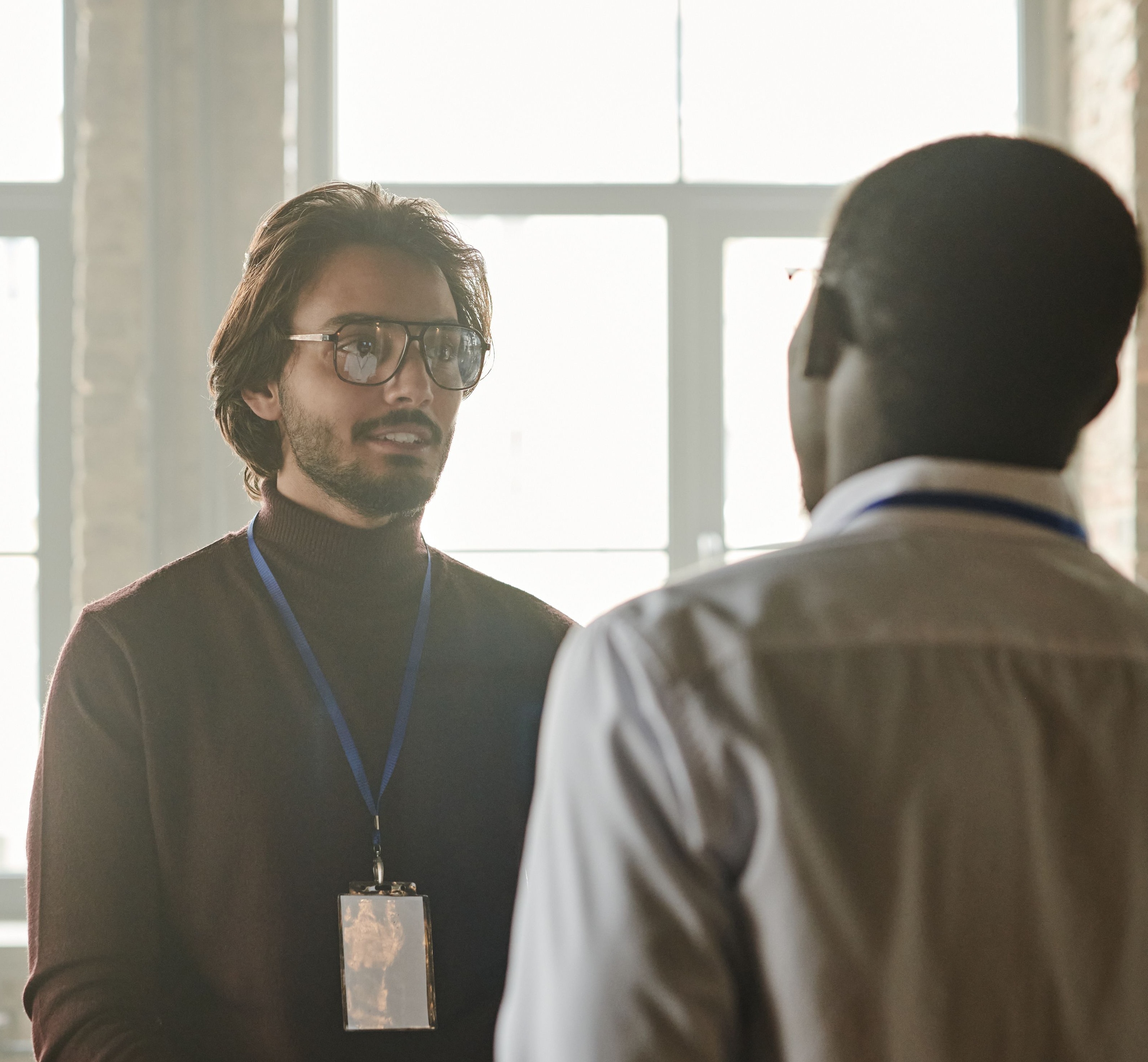 Dois homens conversando em um ambiente de trabalho, um deles a contraluz de uma janela