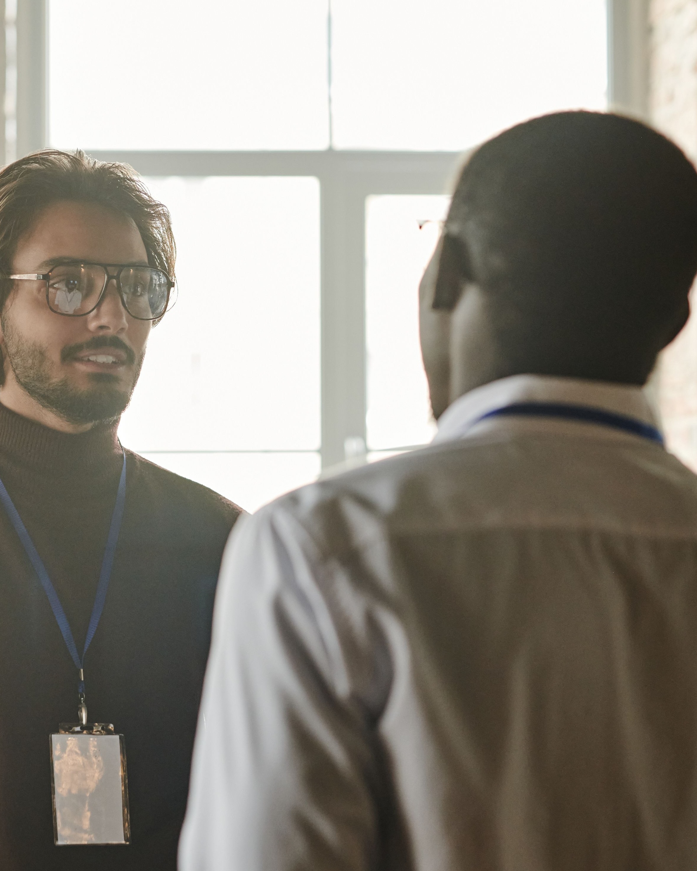 Dois homens conversando em um ambiente de trabalho, um deles a contraluz de uma janela